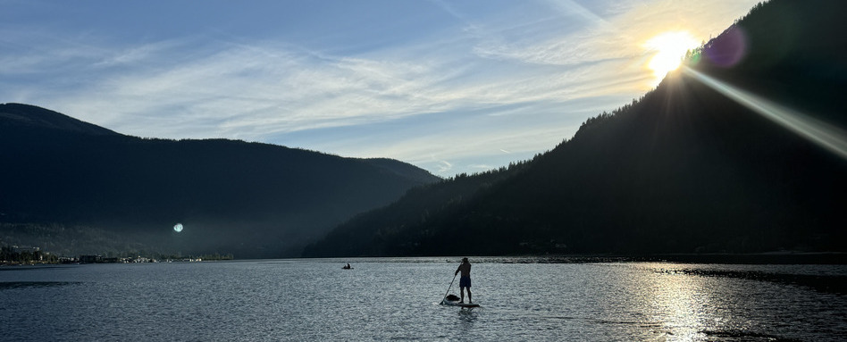 A person paddleboarding in a calm bay with the sun peaking out from behind mountains in the background. A person paddleboarding in a calm bay with the sun peaking out from behind mountains in the background.
