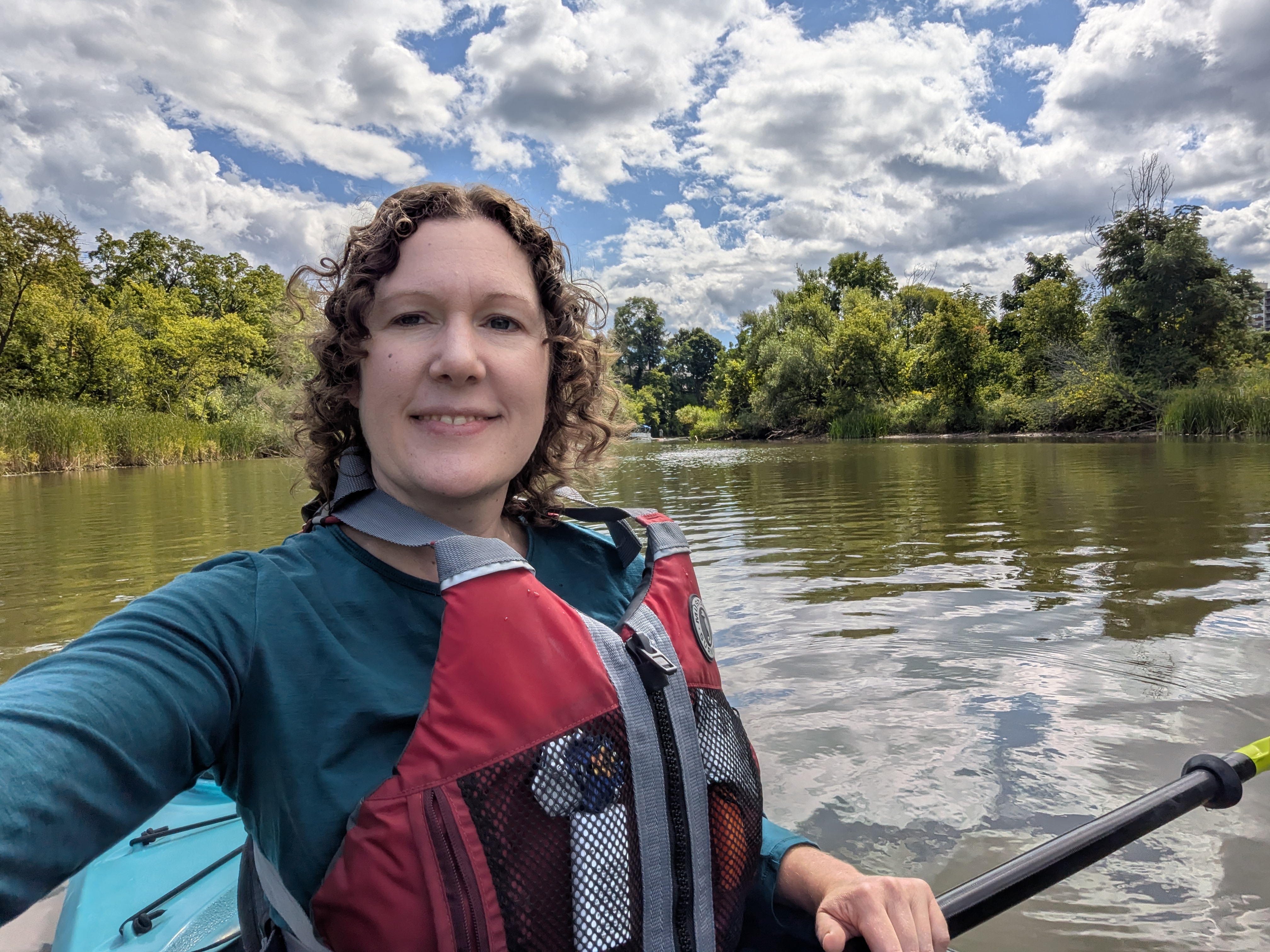 Carolyn takes a selfie in her kayak with water and lush trees in the background. Carolyn takes a selfie in her kayak with water and lush trees in the background.