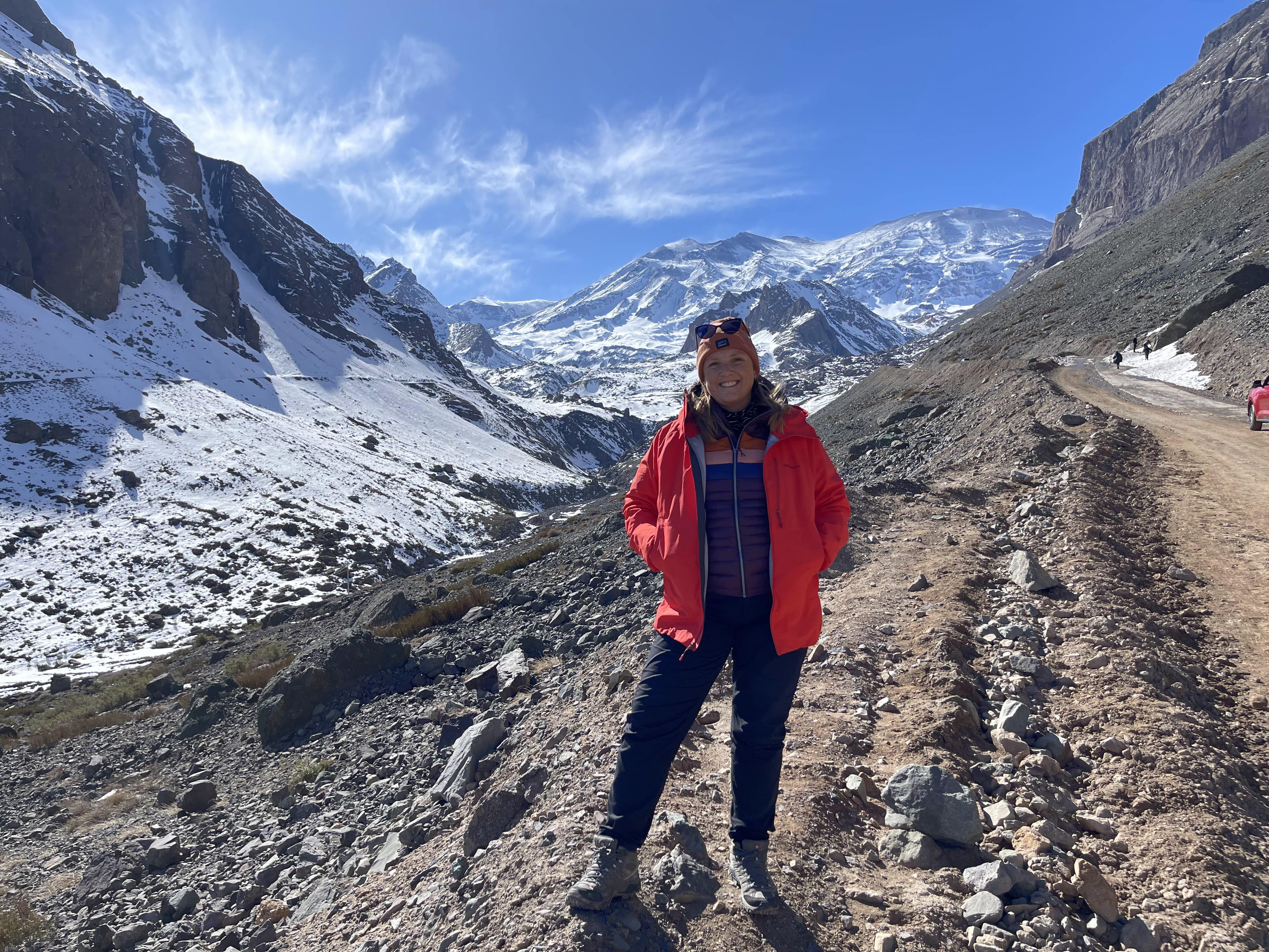 Stacey stands on the trail on a mountain trail with blue sky and snowy peaks in the background. Stacey stands on the trail on a mountain trail with blue sky and snowy peaks in the background.